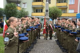 Grupa osób w mundurach stoi w dwóch szeregach, zwróconych ku sobie, składając przysięgę. Na środku pomnik, w tle bloki mieszkalne.