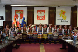 Grupa ludzi w tradycyjnych polskich strojach ludowych stoi na scenie przed flagami Polski, Unii Europejskiej oraz banerami z herbem Polski i napisami.