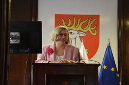 Opis zdjęcia: Kobieta wygłaszająca przemówienie za mównicą z mikrofonem, w tle widoczny herb na ścianie oraz flagi Polski i Unii Europejskiej.