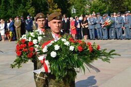 Żołnierze w mundurach niosą wiązanki kwiatów na ceremonii, w tle obserwujący ludzie i inni żołnierze.