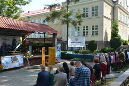 Grupa ludzi siedzi na ławkach i krzesłach na zewnątrz, obserwując uroczystość. Jest scena, namioty i banner z napisem "Powiat Lubelski". Tło to budynek i drzewa.