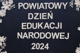 Baner z napisem "Powiatowy Dzień Edukacji Narodowej 2024" z białymi papierowymi kwiatami na granatowym tle.