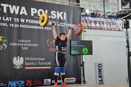 Młody sportowiec wykonuje podrzut sztangi na zawodach. Widoczne 113 kg, tablica wyników pokazuje zielone znaki akceptacji.