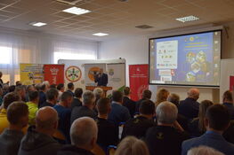 Mężczyzna przemawia na podium przed publicznością w sali konferencyjnej. Na ekranie wyświetlona jest prezentacja, a wokół znajdują się banery promocyjne. Pomieszczenie wypełnione jest słuchaczami.