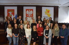 Grupa ludzi pozuje do zdjęcia w sali z drewnianymi panelami. W tle widać trzy plansze: z orłem, herbem oraz mapą powiatu lubartowskiego. Osoby są uśmiechnięte, stoją w kilku rzędach.
