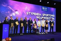 Grupa osób stoi na scenie z dużym banerem w tle o treści "Stypendia Sportowe Województwa Lubelskiego". Każda osoba trzyma teczkę, w środku znajduje się również osoba na wózku inwalidzkim.