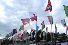 Zbiór różnorodnych flag na wysokich masztach, umieszczonych na stalowej konstrukcji. W tle widać obłoki na niebie oraz zarys drzew.