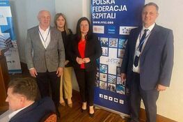 Cztery osoby stoją obok siebie w formalnych strojach na tle baneru Polskiej Federacji Szpitali. W tle drewniana podłoga i biała ściana.