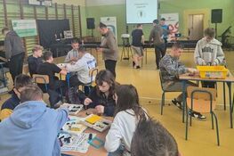 W sali gimnastycznej grupa osób siedzi przy stołach, gdzie pracują nad projektami lub zadaniami. W tle znajduje się ekran projekcyjny oraz sprzęt nagłaśniający. Atmosfera jest edukacyjna i skupiona.