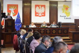 Na zdjęciu widać ludzi siedzących przy stole na tle flag i herbu. Sala jest pełna uczestników, trwa spotkanie lub konferencja. Widoczne są symbole narodowe i regionalne.