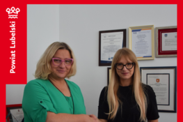 Dwie kobiety podają sobie ręce w biurze, na tle ściany z dyplomami i certyfikatami. Jedna z kobiet trzyma dokumenty związane z rozbudową zakładu opiekuńczo-pielęgnacyjnego w Bełżycach.