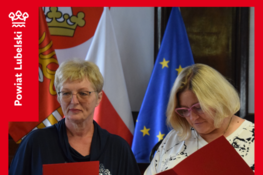Dwie osoby stoją z dokumentami w rękach. W tle widoczne są flagi Polski, Unii Europejskiej i powiatu lubelskiego. Sytuacja wygląda na oficjalne wydarzenie związane z wręczaniem aktów powołania.