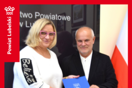 Mężczyzna i kobieta stoją obok siebie. Kobieta trzyma niebieską teczkę z logo, mężczyzna kładzie na niej rękę. W tle flaga Polski i czarne tło z napisem "Starostwo Powiatowe".