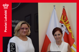 Dwie kobiety pozują do zdjęcia we wnętrzu biura. Jedna z nich trzyma dokument. W tle znajduje się flaga i herb. Plansza informuje o dofinansowaniu dla gminy Bychawa na zakup busa dla osób z niepełnosprawnościami.