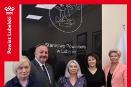 Grupa osób stoi przed szyldem Starostwa Powiatowego w Lublinie. W centrum kobieta trzyma niebieską teczkę. Obok widać flagę Polski. Tekst informuje o dofinansowaniu dla Gminy Bełżyce na zakup busa.