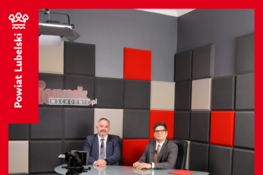 Dwóch mężczyzn siedzi przy okrągłym stole w studiu z szarymi i czerwonymi panelami na ścianie. Przed nimi znajduje się kamera. Na górze napis informuje o wywiadzie dotyczącym dożynek.