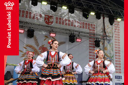 Grupa kobiet w tradycyjnych strojach ludowych tańczy na scenie podczas festiwalu dożynkowego. W tle dekoracje ludowe i logo Powiatu Lubelskiego.