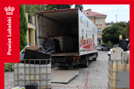Na dziedzińcu przed budynkiem stoi ciężarówka z otwartymi drzwiami, otoczona pojemnikami pełnymi zużytego sprzętu elektronicznego. W tle widoczne są budynki i drzewa. Na czerwonej ramce widnieją napisy informujące o zbiórce.