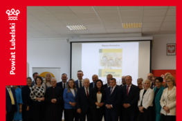 Grupa osób stoi razem w sali, na tle ekranu z prezentacją. Na slajdzie widnieje napis związany z pszczelarstwem. Obok nich stoją flagi i banery z logo "Powiat Lubelski".