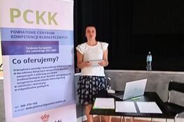 Osoba stoi obok roll-upu z logo "PCKK" i szczegółami dotyczącymi Powiatowego Centrum Kompetencji Klimatycznych. W tle czarne zasłony, stolik z krzesłami i butelka wody.