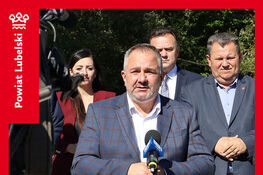 Mężczyzna przemawia na zewnątrz podczas otwarcia drogi i mostu. Wokół niego stoją inni oficjele. Za nim widoczne jest zadrzewione tło. Na górze i dole zdjęcia znajdują się informacje o wydarzeniu.