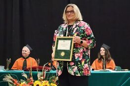 A woman stands holding a framed award, wearing glasses and a floral pattern jacket. In the background, two people in academic robes sit at a table adorned with flowers and documents. The setting appears formal, possibly an academic ceremony.