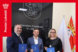 Trzy osoby stojące w biurze trzymają niebieskie teczki. W tle widoczne logo Starostwa Powiatowego w Lublińcu oraz flagi. Na dole napis: "Kolejna umowa w ramach projektu e-Geodezja II".