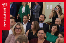 Grupa ludzi siedzi w pomieszczeniu podczas oficjalnego wydarzenia. W tle widoczna jest kobieta fotografująca. Ubrania uczestników są eleganckie. Na czerwonym tle znajdują się teksty informujące o podpisaniu umowy.