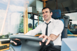 Mężczyzna w białej koszuli prowadzi autobus, patrzy przed siebie, zza pleców widać siedzących pasażerów.