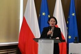 Na zdjęciu wystąpienie kobiety przy mównicy na tle polskiej, unijnej i innej flagi. Obok ekran z napisem o spotkaniu z organizacjami pozarządowymi dotyczącym konsultacji na 2026 rok. Data: 6 października 2023, Warszawa.