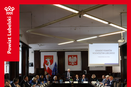 Spotkanie w sali konferencyjnej z dużym stołem w kształcie litery U. Uczestnicy siedzą wokół, na ścianie widać flagi Polski i UE oraz godło Polski. Na ekranie wyświetlony jest napis dotyczący konwentu powiatów.