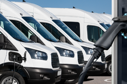 Furgonetki elektryczne w rzędzie, jedna ładowana z ładowarki. Baner programu "NaszEAuto" z logotypami Krajowego Planu Odbudowy, Rzeczpospolitej Polskiej, UE i NFOŚIGW.