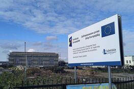 Na zdjęciu znajdują się tablica informacyjna dotycząca dofinansowania Unii Europejskiej oraz plac budowy z widocznym budynkiem w tle. Tablica informuje o projekcie w Lublinie. Niebo jest częściowo zachmurzone.