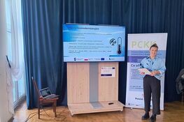 Mężczyzna w niebieskiej koszuli stoi na drewnianej podłodze w sali konferencyjnej, obok dużego ekranu z prezentacją i banneru PCKK. Na stole obok leży otwarty laptop. Zasłony wiszą przy oknie.