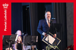 Na scenie mężczyzna przemawia przy mikrofonie, w tle widoczna orkiestra dęta i siedzące dzieci z instrumentami. Obok ustawione są kwiaty. Wydarzenie to jubileusz 30-lecia Gminnej Orkiestry Dętej "Henryczki".