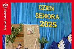 Na scenie udekorowanej niebieskim i czerwonym materiałem siedzi osoba z książką, obok stoją stolik i roślina. Widoczny jest napis "Dzień Seniora 2025".