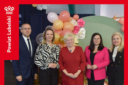 Na zdjęciu pięć osób stoi przed dekoracją z kolorowych balonów. Wszyscy patrzą w stronę aparatu. Osoby są elegancko ubrane. W tle widać elementy wystroju wnętrza.