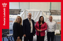 Cztery osoby stoją przed białym namiotem oznaczonym jako "Discovery Space Station". Osoby są elegancko ubrane, pozują do zdjęcia. W tle widać panele słoneczne i niebieską podłogę.