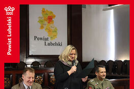 Na zdjęciu trzy osoby siedzą przy stole w sali konferencyjnej. Kobieta stoi z mikrofonem. Z tyłu mapa Powiatu Lubelskiego i herb. Obok wojskowy i mężczyzna w marynarce.