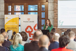 Na zdjęciu widać kobietę przemawiającą na konferencji w sali. Stoi przy mównicy z mikrofonem. Za nią widoczne są banery reklamowe i ekran z prezentacją, a przed nią siedzący uczestnicy.