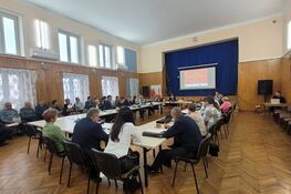 Na zdjęciu widać dużą salę z drewnianą podłogą, wypełnioną ludźmi siedzącymi przy prostokątnych stołach. Na ścianie wisi ekran z prezentacją, a w tle powiewa flaga.