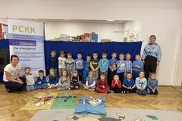 Na zdjęciu grupa dzieci wraz z dwiema dorosłymi osobami stoi w pomieszczeniu. Za nimi znajduje się niebieskie tło oraz baner PCKK. Na podłodze leżą różne przedmioty. Dzieci są ubrane głównie w niebieskie ubrania.
