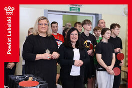 Grupa osób stojąca w sali gimnastycznej podczas turnieju tenisa stołowego. Na pierwszym planie dwie kobiety trzymają trofea. W tle uczestnicy z rakietkami.