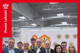 Grupa ludzi stoi razem na tle ściany z logo wydarzenia. Wszyscy są elegancko ubrani i uśmiechają się do zdjęcia. Obok, czerwony pasek z napisem "Pierwszy Dzień Targów Edukacyjnych".