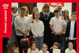 Grupa dzieci i dorosłych pozuje z dyplomami na uroczystości rozstrzygnięcia konkursu „Ozdoba Wielkanocna”. Wszyscy są uśmiechnięci, a na ścianie za nimi widnieje herb powiatu lubelskiego.