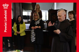 Osoby zgromadzone wokół stołu na wielkanocnym spotkaniu. Na stole znajdują się talerze z przekąskami. Mężczyzna w habicie czyta z notatek. W tle widoczna dekoracja wielkanocna.