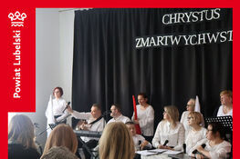 Grupa osób siedzi na scenie podczas występu w Domu Pomocy Społecznej. W tle napis "Chrystus Zmartwychwstał". Większość ubrana na biało. Na pierwszym planie tablica z napisem "Spotkanie Wielkanocne".
