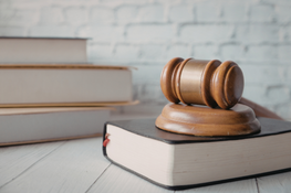 Drewniany młotek sędziowski leży na dużej książce w tle stosu innych książek. Scena na drewnianym stole z białą ceglaną ścianą w tle, sugerująca powiązanie z prawem lub wymiarem sprawiedliwości.