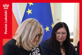 Dwie kobiety siedzą przy stole, podpisując dokumenty. W tle widoczne są flagi Polski i Unii Europejskiej. Na dole znajduje się informacja o podpisaniu umów na inwestycje drogowe w Powiecie Lubelskim.