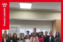 Grupa ludzi stoi uśmiechnięta w nowoczesnej pracowni fryzjerskiej. W tle widać produkty do pielęgnacji włosów i plakat. Dwóch uczestników trzyma duże obrazy.